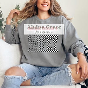 May include: A gray sweatshirt with the word "WIFEY" in a black and white checkered pattern. The text "Alaina Grace Handmade" is above the word "WIFEY". The sweatshirt is worn with blue jeans.