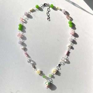 Puede incluir: Un delicado collar de cadena plateada con una variedad de cuentas de colores pastel, incluyendo rosa, verde, blanco y transparente. El collar presenta un pequeño dije de flor blanca y un dije de fresa al final.