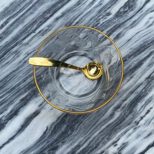 Puede incluir: Un tazón de vidrio transparente con un borde dorado y un diseño floral. Una pequeña cuchara dorada está dentro del tazón.