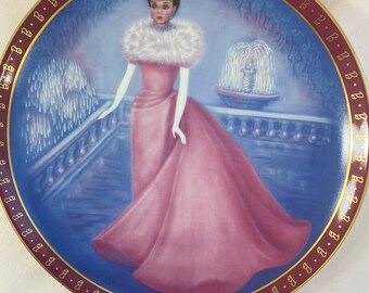 May include: A porcelain plate with a gold rim and a blue background featuring a woman in a pink gown and white gloves standing in front of a fountain. The plate is decorated with a gold border with the letter "B" repeated around the edge.