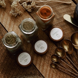 Pode incluir: Três frascos de especiarias quadrados de vidro com tampas de madeira escura, rotulados como Italian Seasoning, Black Pepper e Paprika. Os frascos contêm ervas e especiarias secas. Um conjunto de colheres medidoras de metal dourado está ao lado.