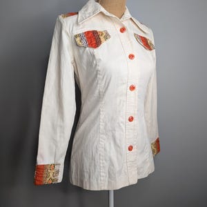 May include: Cream-coloured button-down shirt with long sleeves. The shirt features orange buttons and decorative patches on the chest pockets and cuffs. The patches have a colourful, patterned design. The shirt is displayed on a mannequin.