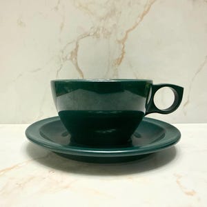 May include: A dark green teacup and saucer set. The cup has a distinctive handle with a circular design. The saucer is round and flat. The set is ceramic with a glossy finish. The set is on a white marble surface.