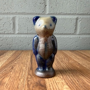 May include: A ceramic bear figurine featuring a speckled glaze with a gradient of cream, brown, and deep blue tones. The bear has a rounded head, small ears, and a smooth, glossy finish, standing upright on a circular base with a distinct vertical line.