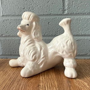 May include: A white ceramic poodle figurine in a reclining pose. The poodle has a detailed, textured coat, a pompadour hairstyle, and a curled tail. The figurine is on a wooden surface with a blue brick background.