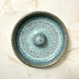 May include: A handmade ceramic soap dish with a speckled, light blue glaze. The dish has a raised center and a dark blue rim. The speckled pattern adds a unique touch to this functional piece. The dish is round and measures approximately 4 inches in diameter.