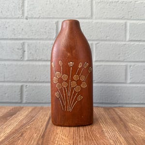 May include: A brown, handcrafted vase with a narrow neck and a rounded body. The vase features an embossed floral design in a lighter brown color. The vase is displayed on a wooden surface against a white brick wall.