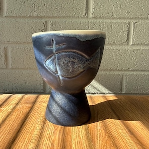 May include: A dark blue ceramic chalice with a flared top and a pedestal base. The chalice features an embossed fish and cross symbol. The rim is a light beige colour. The chalice is sitting on a wooden surface.