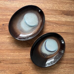 May include: Two oval-shaped ceramic dishes featuring a dark brown, glossy outer rim that transitions into a lighter, speckled blue-grey center. The glaze has a smooth, reflective finish with subtle variations in color depth across the surface.