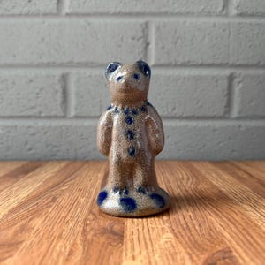 May include: A small, standing bear figurine crafted from speckled tan ceramic. The bear features dark blue painted accents on its ears, eyes, nose, and a vertical row of dots down its chest, with additional blue spots decorating its circular base.
