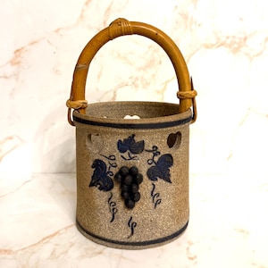 May include: A tan ceramic wine bottle holder with a woven handle. The holder features a dark blue grape and leaf design. The top rim has a dark blue band and heart-shaped cutouts.