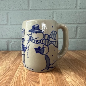 May include: A beige ceramic mug featuring a blue hand-painted design of a snowman wearing a top hat and a striped scarf. The snowman holds a broom, and the mug has a sturdy handle on the right side, with a smooth, glossy finish throughout.