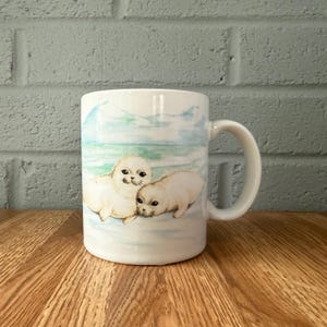 May include: A white ceramic mug featuring a watercolor illustration of two light brown baby seals resting on a light blue and white icy landscape. The mug has a curved handle on the right side and a smooth, glossy finish throughout the exterior surface.