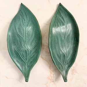 May include: Two teal-colored leaf-shaped ceramic dishes. Each dish features a detailed vein pattern and a slightly raised edge. The dishes are designed to resemble large leaves, perfect for serving or display.