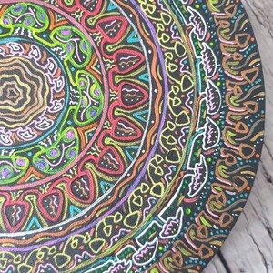 Pode incluir: Um design circular de mandala pintado em uma superf&iacute;cie preta. O mandala apresenta padr&otilde;es intrincados em tons de rosa, verde, amarelo, azul e vermelho.