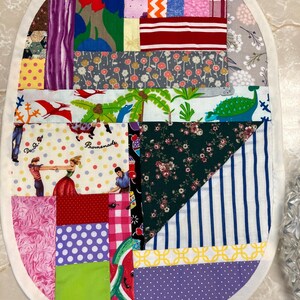 May include: An oval-shaped fabric mat featuring a patchwork design of various colourful patterns, including polka dots, stripes, floral prints, and a scene with dancing figures and dinosaurs, all bordered by a solid white fabric trim.