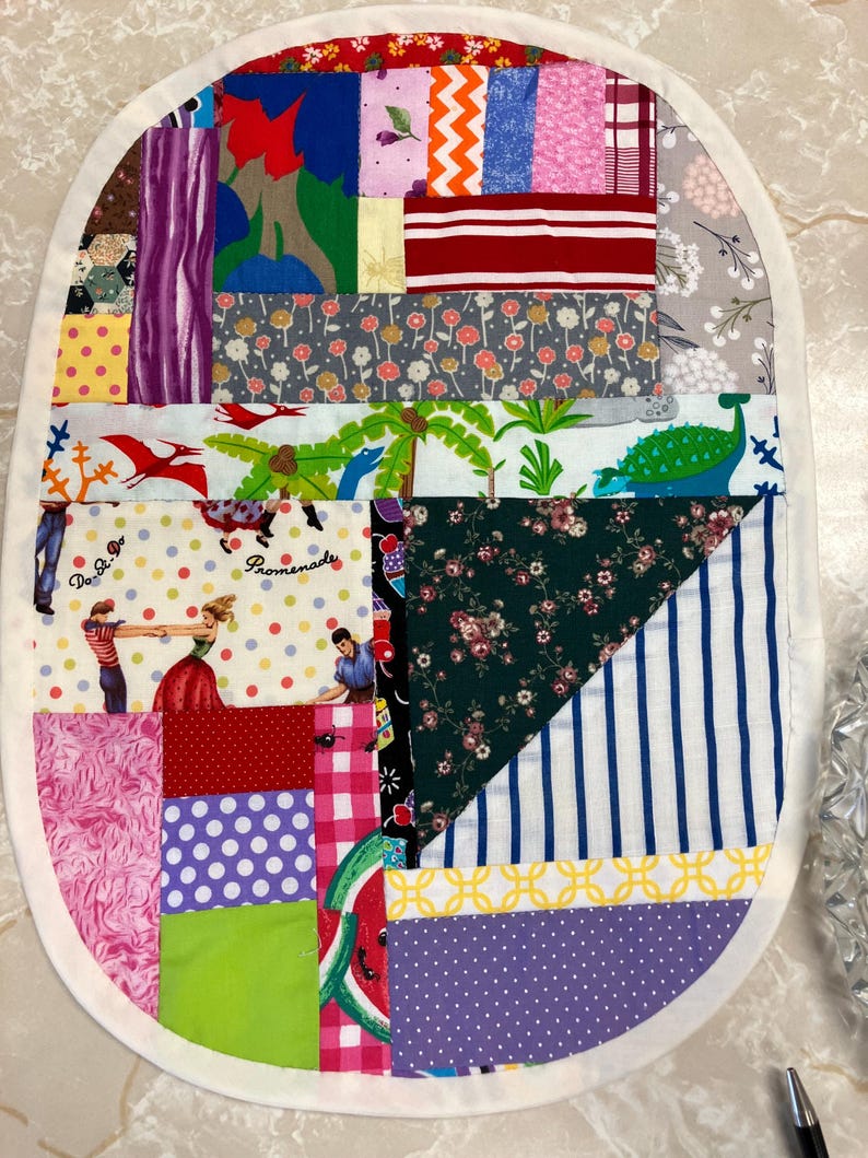 May include: An oval-shaped fabric mat featuring a patchwork design of various colourful patterns, including polka dots, stripes, floral prints, and a scene with dancing figures and dinosaurs, all bordered by a solid white fabric trim.