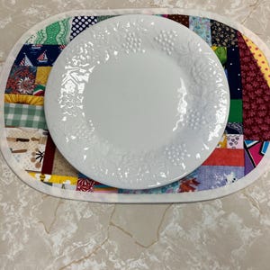 May include: A white ceramic plate with a raised grape and leaf pattern sits centred on an oval fabric placemat. The placemat features a patchwork design composed of various colourful cotton fabric squares with diverse patterns and prints.