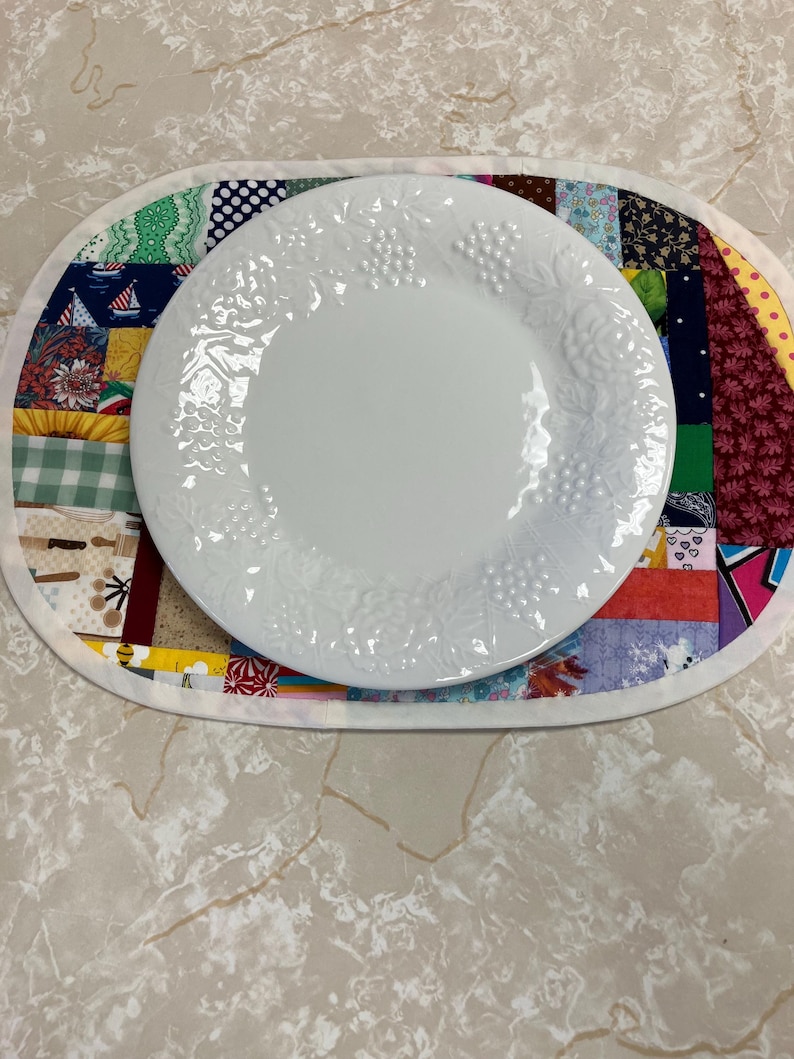 May include: A white ceramic plate with a raised grape and leaf pattern sits centred on an oval fabric placemat. The placemat features a patchwork design composed of various colourful cotton fabric squares with diverse patterns and prints.