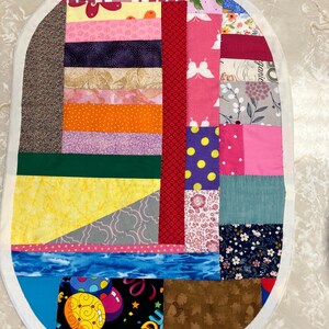May include: An oval-shaped fabric mat featuring a patchwork design of various colourful cotton textiles. The quilt-style piece includes patterns of butterflies, flowers, polka dots, and geometric shapes in shades of red, yellow, blue, and pink.
