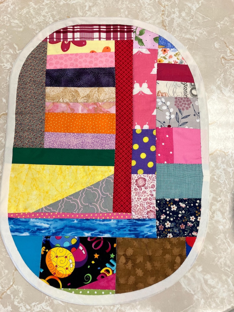 May include: An oval-shaped fabric mat featuring a patchwork design of various colourful cotton textiles. The quilt-style piece includes patterns of butterflies, flowers, polka dots, and geometric shapes in shades of red, yellow, blue, and pink.