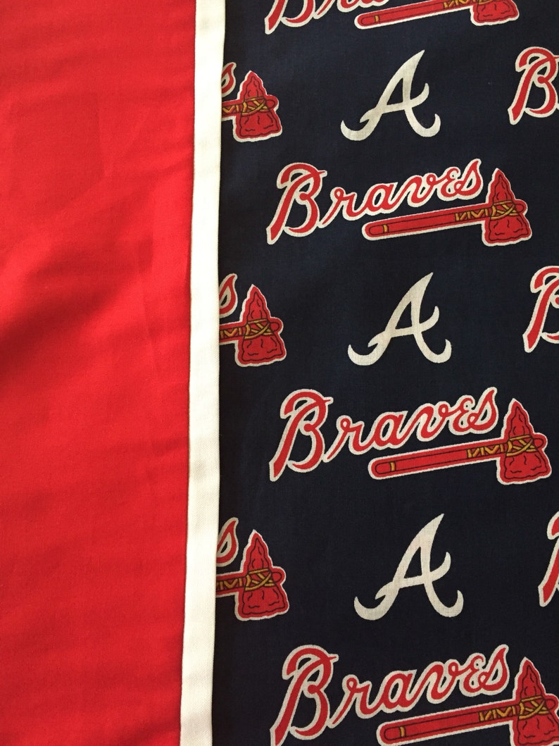 May include: A red and navy blue fabric with a white border. The navy blue side features the word "Braves" in red script, along with a red tomahawk and the letter "A".