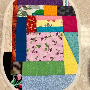 May include: An oval-shaped fabric mat constructed from various multicoloured patchwork quilt squares. The design features diverse patterns including floral prints, geometric shapes, polka dots, and solid colours, all bordered by a white fabric trim.