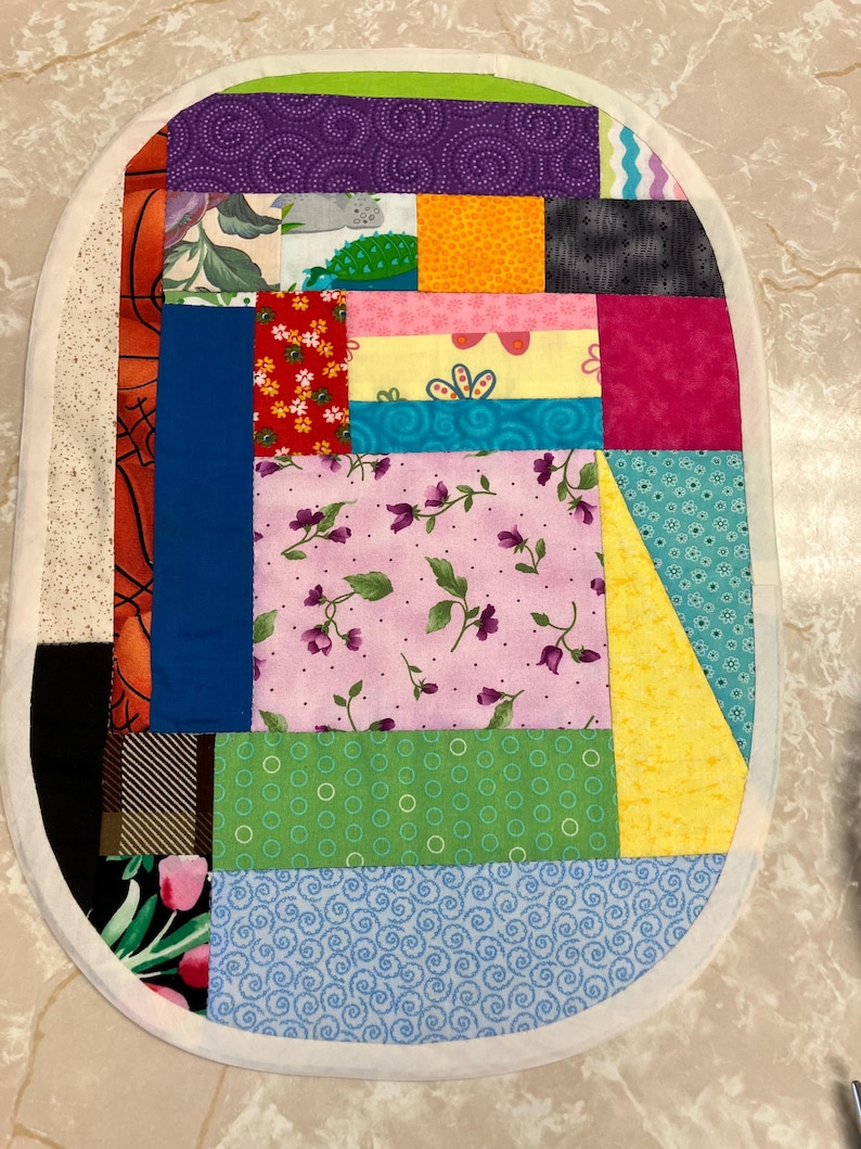 May include: An oval-shaped fabric mat constructed from various multicoloured patchwork quilt squares. The design features diverse patterns including floral prints, geometric shapes, polka dots, and solid colours, all bordered by a white fabric trim.