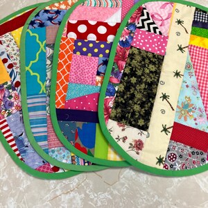 May include: A set of four oval-shaped fabric placemats featuring a patchwork design of various colorful patterns including polka dots, floral prints, stripes, and palm trees. Each placemat is finished with a bright green fabric border trim.