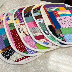 May include: A set of five oval-shaped fabric placemats featuring a patchwork design with various colorful patterns, including floral, polka dot, plaid, and geometric prints. Each placemat is finished with a solid white fabric border trim.