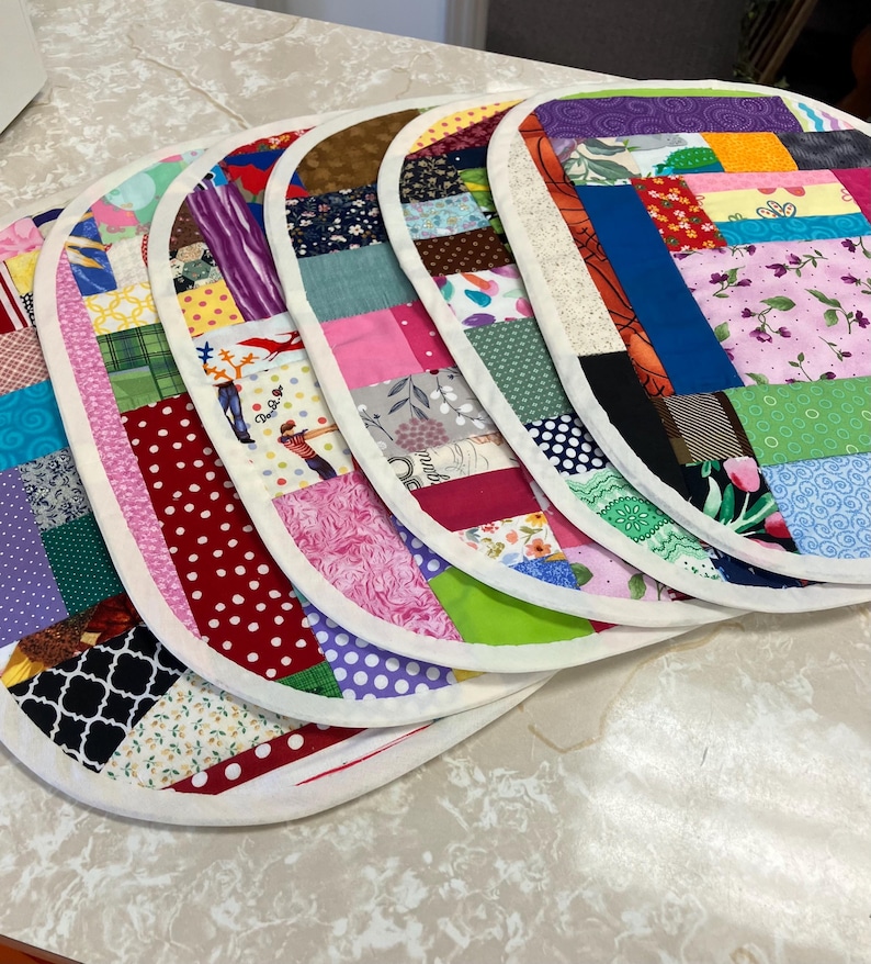 May include: A set of five oval-shaped fabric placemats featuring a patchwork design with various colourful patterns, including floral, polka dot, plaid, and geometric prints. Each placemat is finished with a solid white fabric border trim.