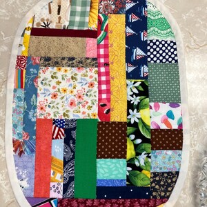 May include: An oval-shaped fabric placemat featuring a patchwork design composed of various rectangular fabric scraps in diverse colours and patterns, including floral, gingham, polka dot, nautical, and fruit prints, finished with a white border.
