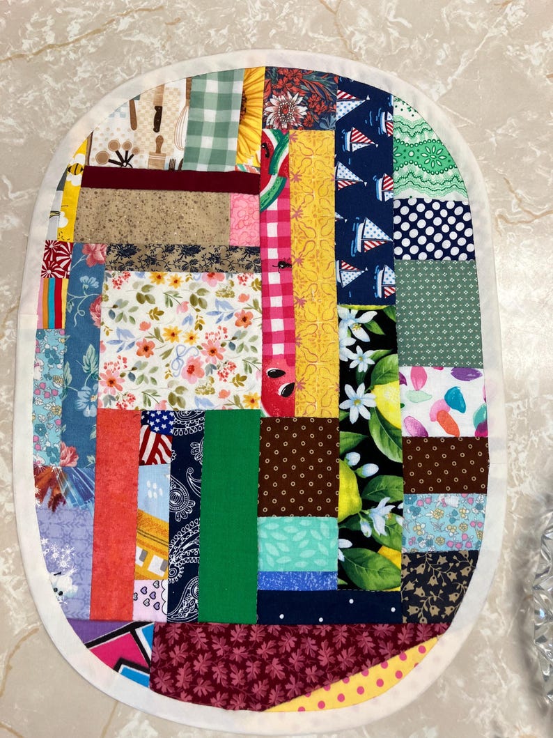May include: An oval-shaped fabric placemat featuring a patchwork design composed of various rectangular fabric scraps in diverse colours and patterns, including floral, gingham, polka dot, nautical, and fruit prints, finished with a white border.