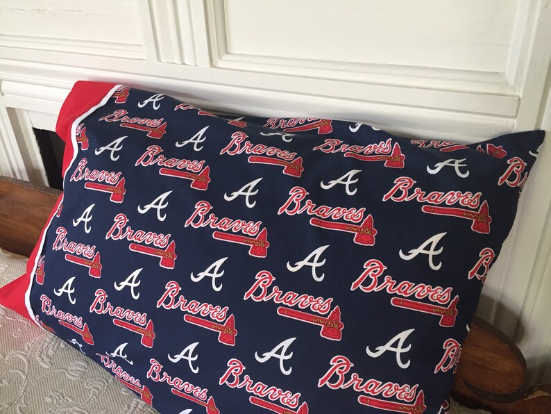 May include: A navy blue pillowcase featuring a repeating pattern of the Atlanta Braves logo and wordmark in red and white. The pillowcase has a red border.