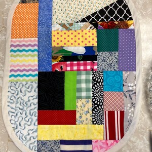 May include: An oval-shaped patchwork mat constructed from various fabric scraps featuring a wide array of colours and patterns, including polka dots, stripes, floral prints, and geometric shapes. The mat is finished with a solid cream-coloured border.