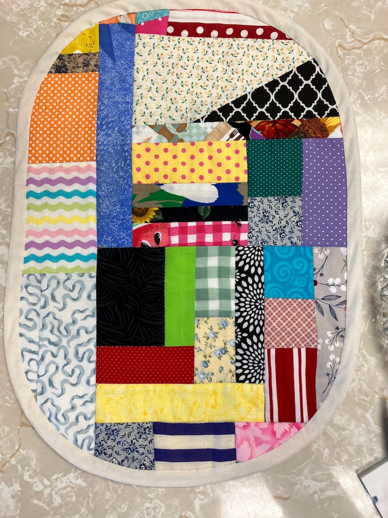 May include: An oval-shaped patchwork mat constructed from various fabric scraps featuring a wide array of colours and patterns, including polka dots, stripes, floral prints, and geometric shapes. The mat is finished with a solid cream-coloured border.