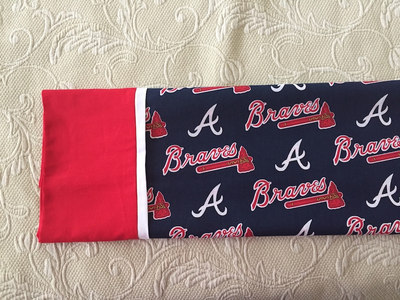 May include: A pillowcase featuring the Atlanta Braves logo and team name. The design includes the letter "A" and the word "Braves" in white script, along with a tomahawk logo, set against a navy blue background. The pillowcase has a red border.
