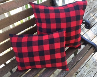 May include: Two red and black plaid throw pillows on a wooden bench. The pillows are made of a soft, textured fabric and have a classic plaid pattern.