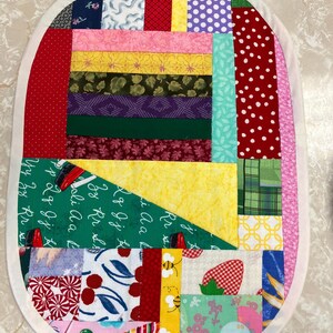 May include: An oval-shaped patchwork quilt mat featuring a variety of colourful fabric scraps including red polka dots, yellow, green, and floral patterns. A dark green section displays white cursive alphabet letters. The border is a solid cream fabric.