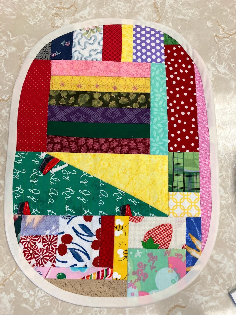 May include: An oval-shaped patchwork quilt mat featuring a variety of colourful fabric scraps including red polka dots, yellow, green, and floral patterns. A dark green section displays white cursive alphabet letters. The border is a solid cream fabric.