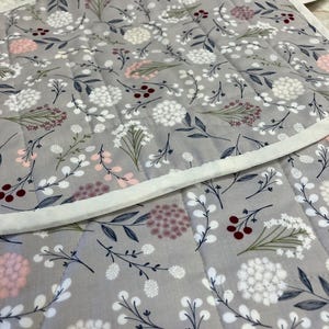 May include: A quilted fabric mat featuring a light grey background with a repeating pattern of white, pink, and dark red floral sprigs and berries. The mat has a white fabric border and straight quilted stitching lines across the surface.