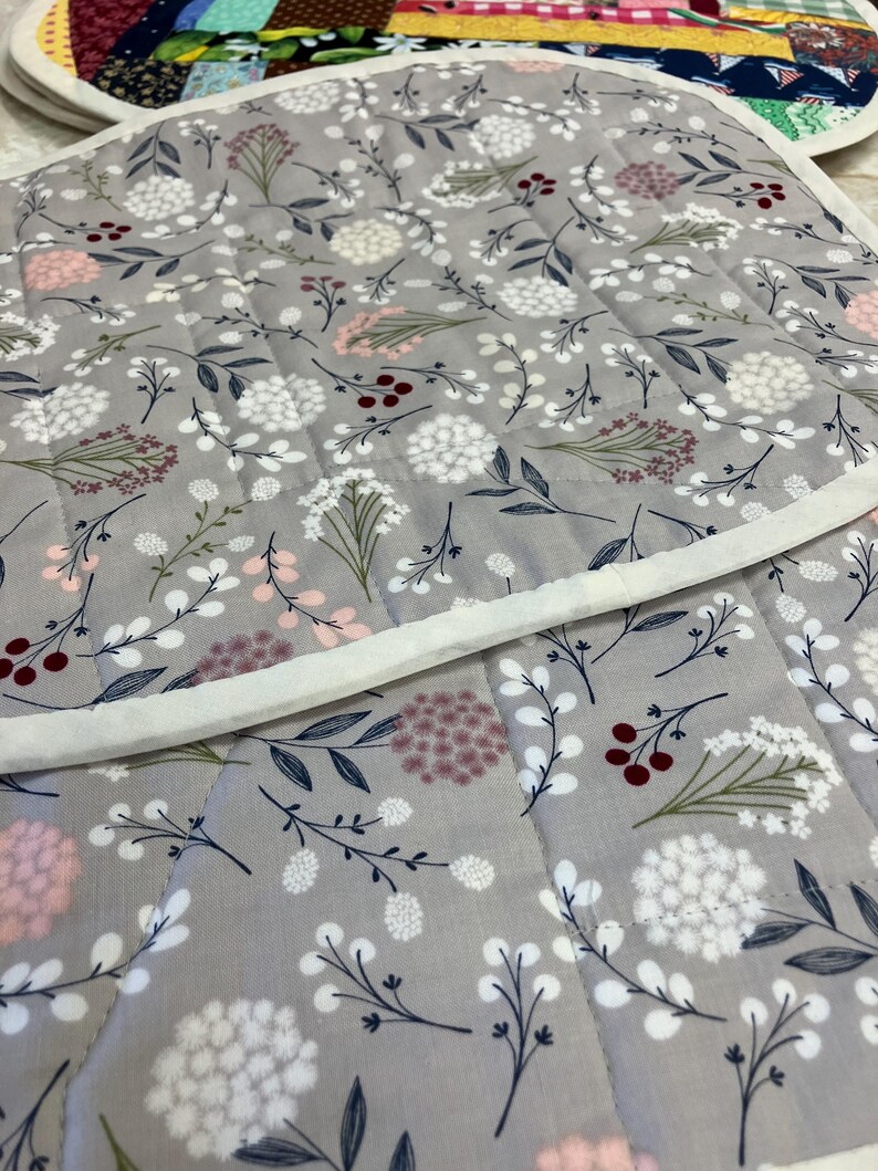 May include: A quilted fabric mat featuring a light grey background with a repeating pattern of white, pink, and dark red floral sprigs and berries. The mat has a white fabric border and straight quilted stitching lines across the surface.