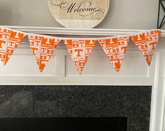 May include: A pennant banner featuring orange and white triangular flags. Each flag displays the University of Tennessee logo. The banner is strung across a white surface, with a wooden sign that says "Welcome" in the background.