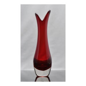 May include: A tall, slender glass vase featuring a deep red translucent body that transitions into a clear, thick glass base. The top opening of the vase has a distinct V-shaped cutout, creating two upward-pointing peaks on either side of the rim.