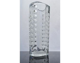 Peut inclure: Un vase en verre transparent avec une surface texturée. Le vase a une forme rectangulaire avec des bords arrondis et une large ouverture en haut.