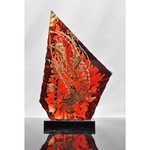 May include: A triangular, translucent amber-colored resin sculpture containing preserved marine life. Inside the resin are two seahorses, a crab, and white coral branches. The sculpture is mounted on a rectangular black base.