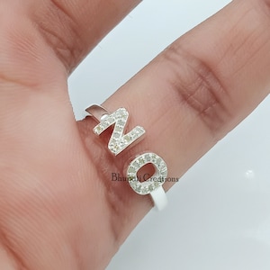 Puede incluir: Un anillo de plata con las letras "NO" engastadas con pequeñas piedras transparentes. El anillo está en un dedo.