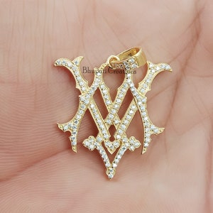 May include: Gold pendant with a diamond-encrusted design featuring the letters "M" and "V" intertwined. The pendant is on a white background.