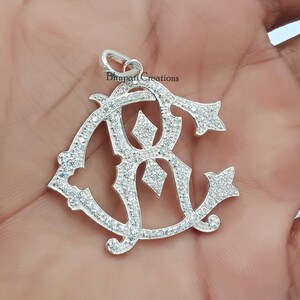 May include: Silver pendant with a detailed design of the letters "RC" set with clear cubic zirconia stones. The pendant has a loop at the top for attaching to a chain.