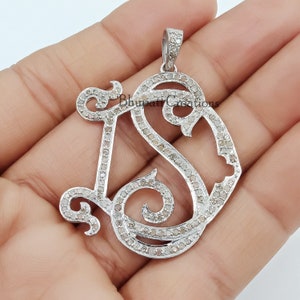 May include: A silver pendant with a monogram design featuring the letters 'IS' set with small diamonds. The pendant is on a hand.